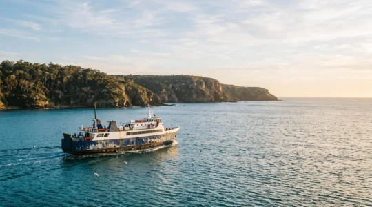 A red-hulled Australian ferry crossing calm turquoise coastal waters near a rugged island coastline at golden hour