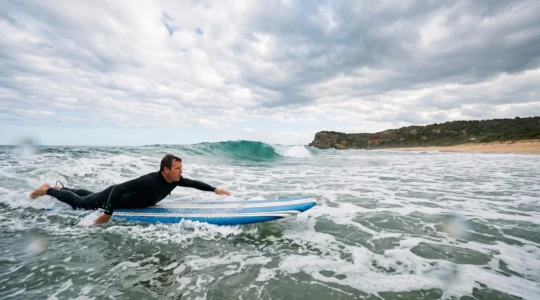 A surfer paddling from calm whitewater toward a rolling green wave under an open Australian sky, symbolizing progression from beginner to intermediate skill.