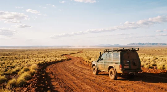 A solitary 4WD vehicle crossing a vast red desert track under an enormous open sky, symbolizing the remoteness of the Australian Outback
