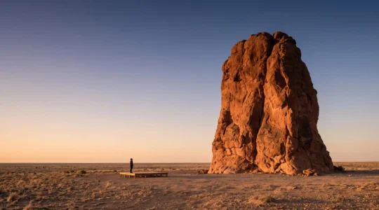 Wide dawn view of a massive red sandstone monolith rising alone from the flat desert plain, symbolizing Uluru's geological scale and cultural sacredness