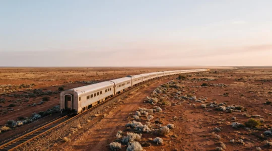 A long silver passenger train crossing the vast, empty red-ochre expanse of the Australian outback under a pale sky