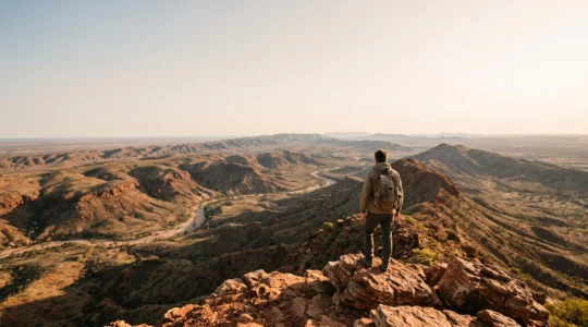 A lone hiker with a loaded backpack standing on a rocky ridge overlooking a vast red-earth Australian outback landscape at golden hour