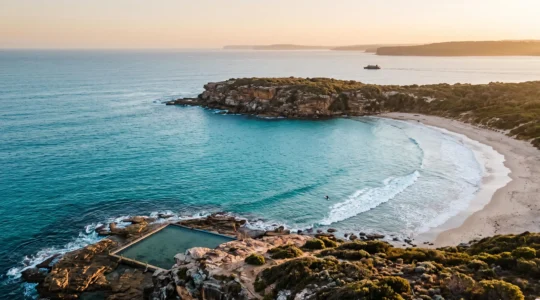 Wide aerial view of an Australian turquoise bay at golden hour, showing a surfer paddling out near a rocky headland and an ocean pool in the foreground